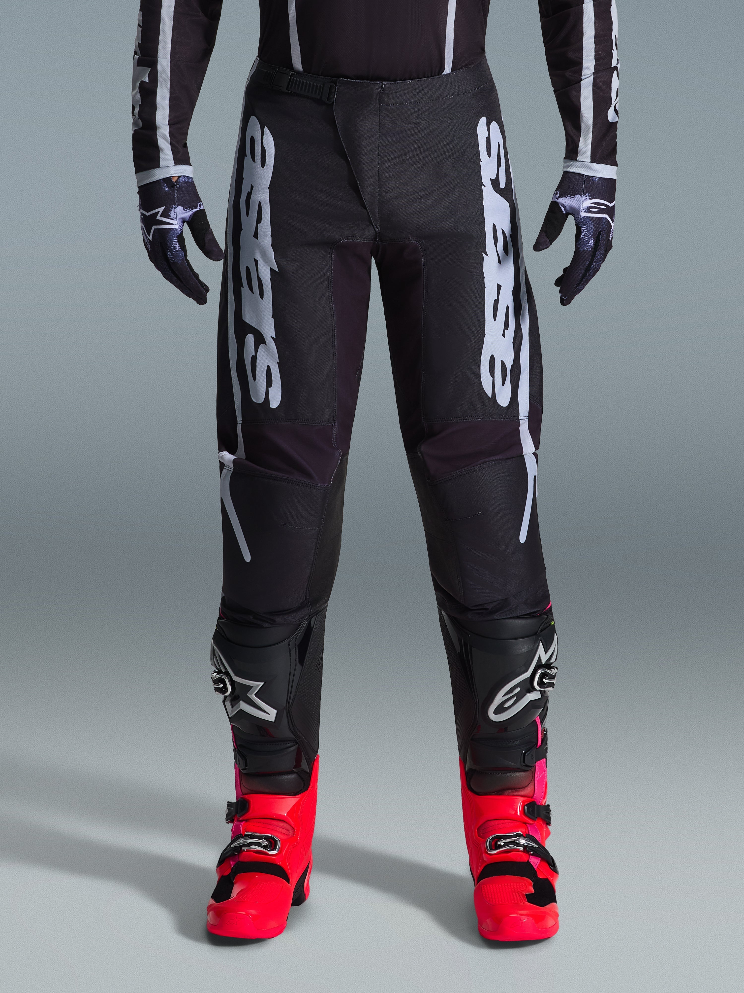 Alpinestars 2026 Fluid Apex Pants, pantalon de motocross noir avec un grand lettrage blanc, porté avec des protections de genoux noires et des bottes rouge vif, châssis léger en poly-tissu 600D, design sport pour la course tout-terrain