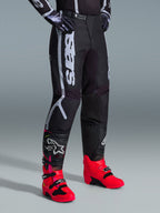 Pantalon Alpinestars Fluid Apex 2026, pantalon de motocross, noir et gris, porté avec des bottes rose vif et rouges et un maillot noir et blanc assorti, doté d'un marquage Alpinestars vertical sur la cuisse, de panneaux renforcés et d'une fermeture à cliquet à la taille pour une performance tout-terrain.