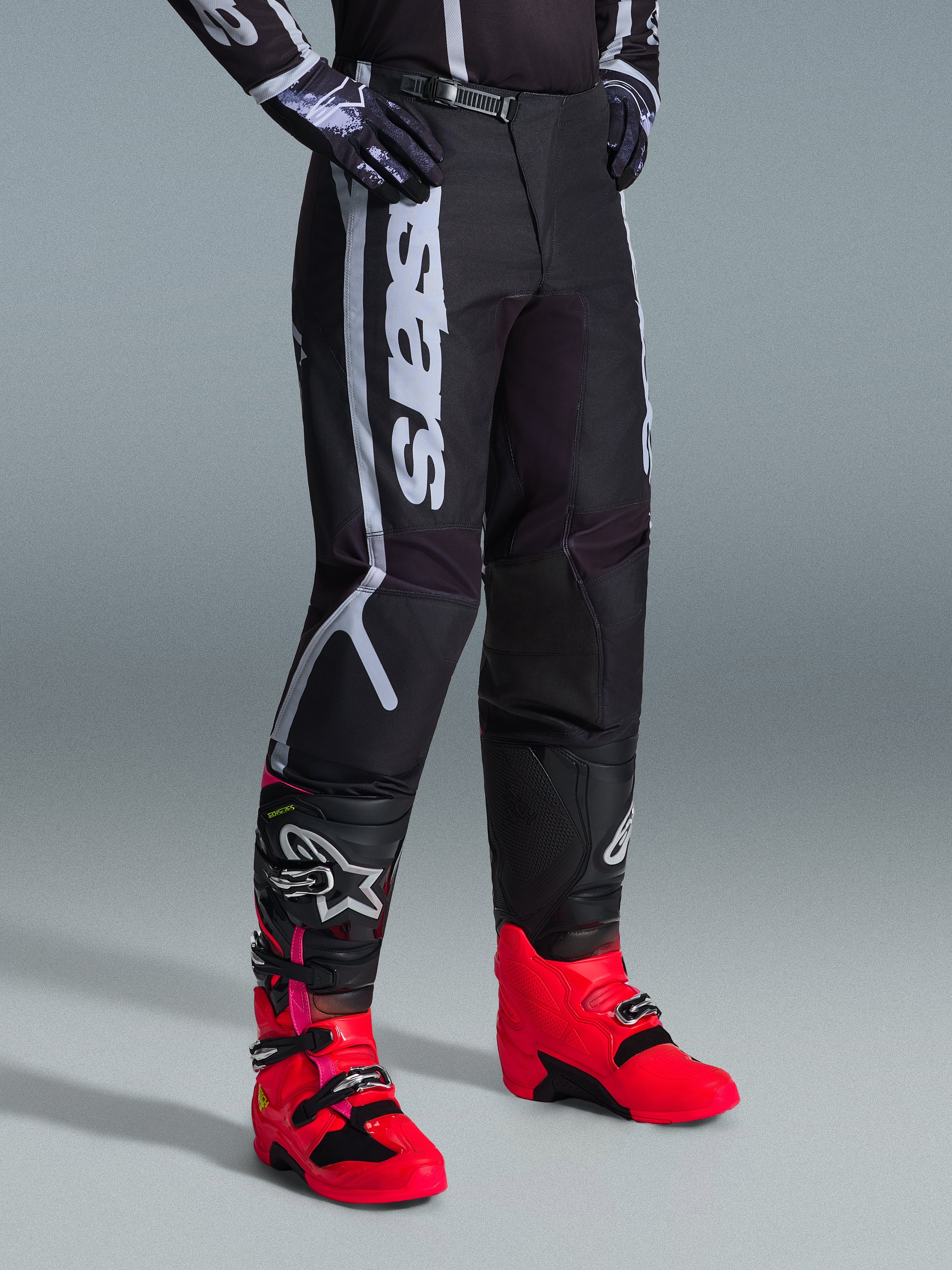 Pantalon Alpinestars Fluid Apex 2026, pantalon de motocross, noir et gris, porté avec des bottes rose vif et rouges et un maillot noir et blanc assorti, doté d'un marquage Alpinestars vertical sur la cuisse, de panneaux renforcés et d'une fermeture à cliquet à la taille pour une performance tout-terrain.