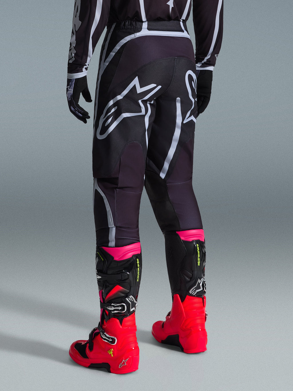 Alpinestars Fluid Apex Pants 2026, pantalon de motocross, noir et gris, vue arrière présentant de grands logos Alpinestars blancs sur l'assise, porté avec des bottes rouge vif et rose, construction durable en poly-tissu 600D pour des performances tout-terrain