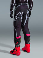 Alpinestars Fluid Apex Pants 2026, pantalon de motocross, noir et gris, vue arrière présentant de grands logos Alpinestars blancs sur l'assise, porté avec des bottes rouge vif et rose, construction durable en poly-tissu 600D pour des performances tout-terrain