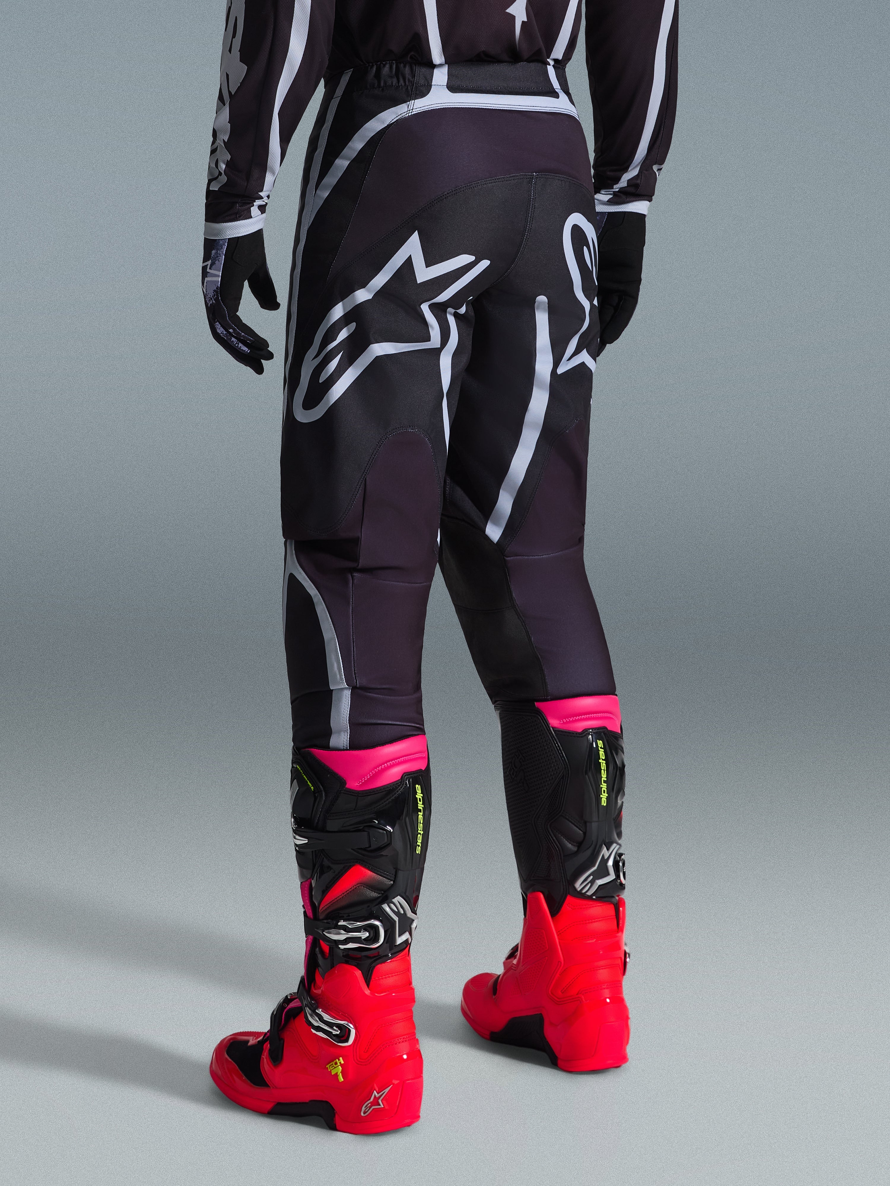 Alpinestars Fluid Apex Pants 2026, pantalon de motocross, noir et gris, vue arrière présentant de grands logos Alpinestars blancs sur l'assise, porté avec des bottes rouge vif et rose, construction durable en poly-tissu 600D pour des performances tout-terrain