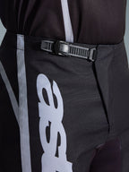Pantalon Alpinestars Fluid Apex 2026, pantalon de motocross, noir et gris, gros plan sur le système de fermeture à cliquet intégré pour un ajustement sécurisé, avec graphismes de logo blancs sur un poly-tissu 600D durable