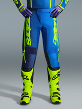 Alpinestars 2026 Fluid Apex Pants, pantalon de motocross, bleu et jaune fluo, porté avec des bottes jaune fluo assorties et des gants violets, logo jaune vif sur les côtés, design ergonomique pour la course tout-terrain