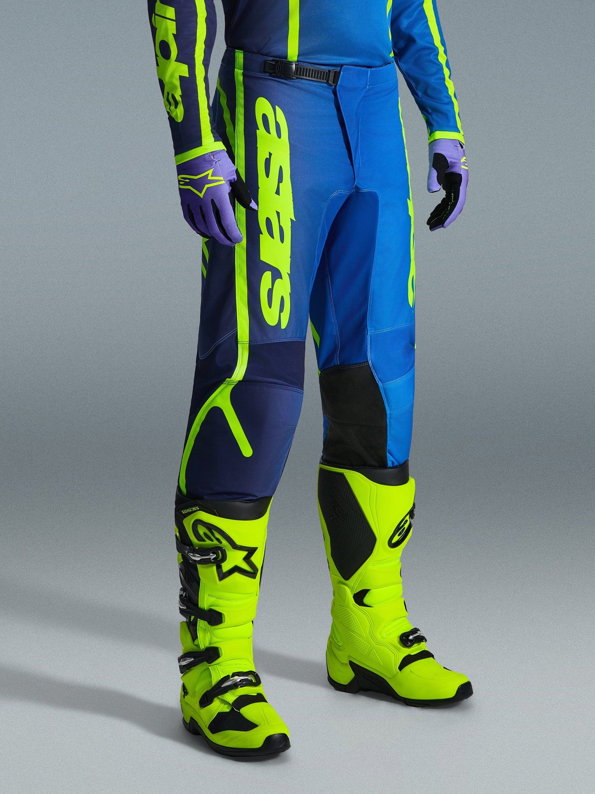 Pantalon Alpinestars Fluid Apex 2026, pantalon de motocross, bleu et jaune fluo, porté avec des gants assortis et des bottes jaune fluo éclatant, doté d'un marquage vertical audacieux, de panneaux de genoux renforcés et d'une fermeture à cliquet sécurisée à la taille