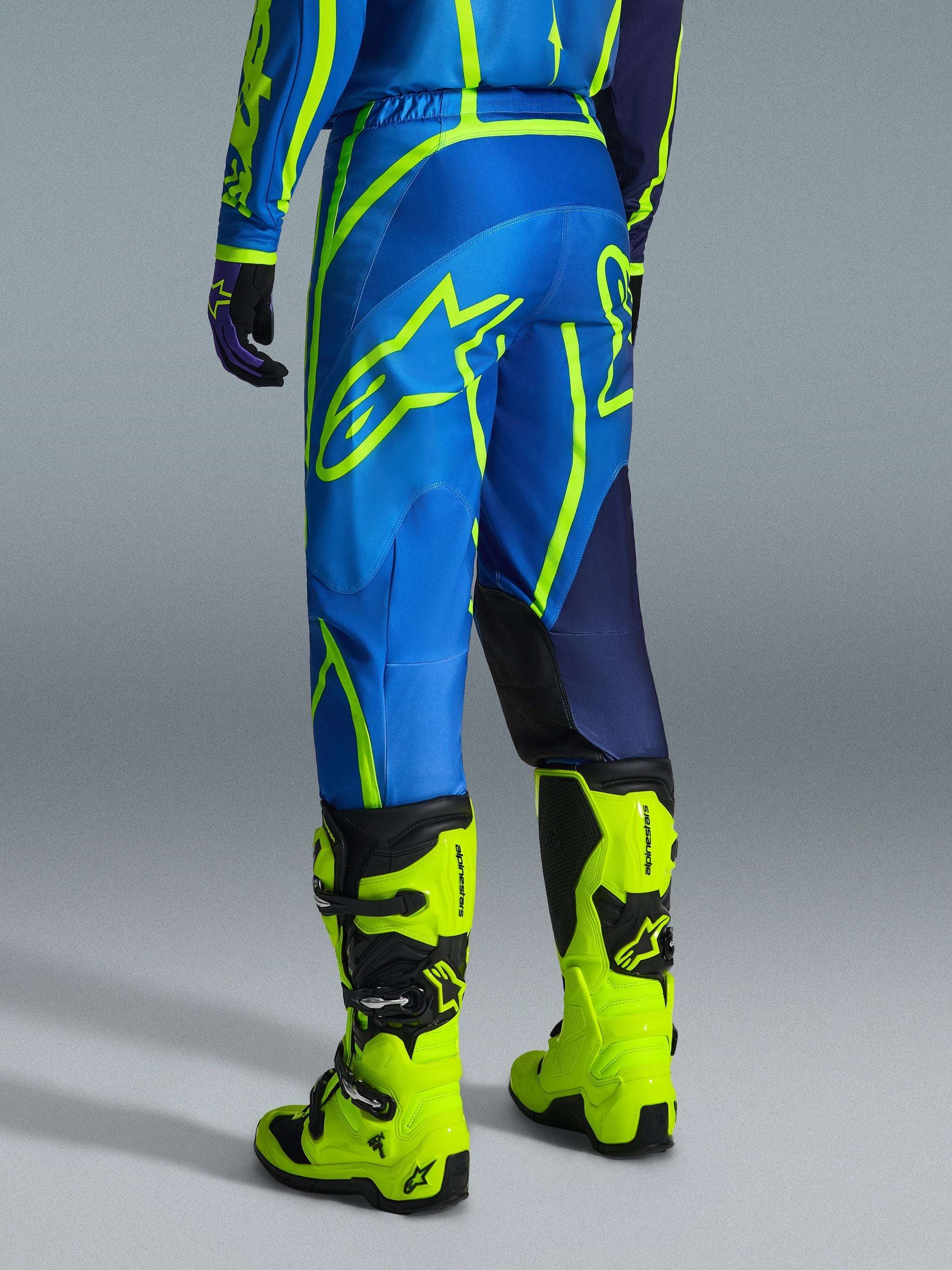 Alpinestars 2026 Fluid Apex Pants, pantalon de motocross, bleu et jaune fluo, vue arrière d'un pilote portant une tenue assortie et des bottes jaune fluo, arborant des logos Alpinestars audacieux et un design ergonomique