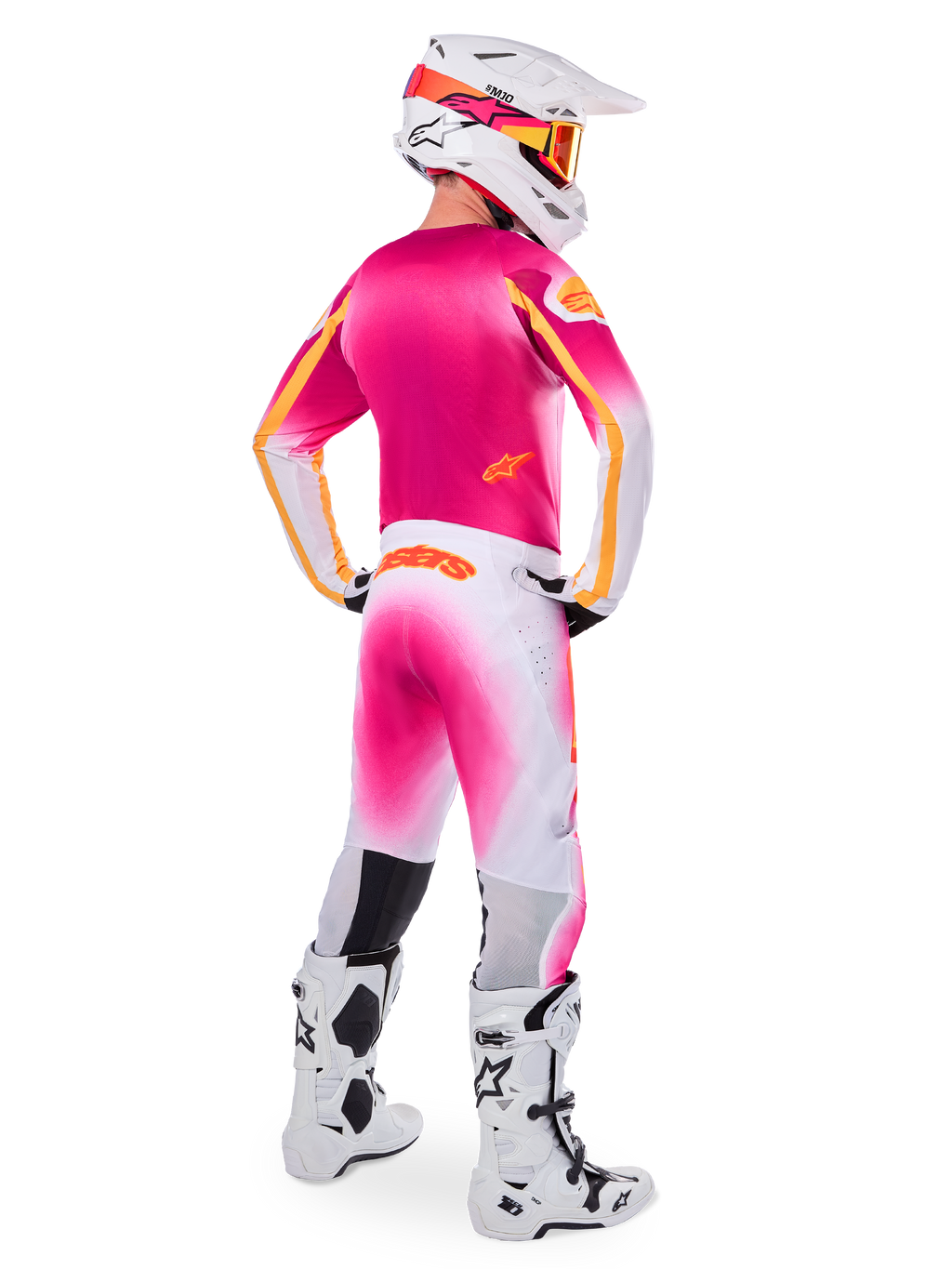 Pilote de motocross vu de dos portant la tenue Limited Edition Supertech AFD MIG, maillot rose avec accents orange, pantalon blanc avec dégradé rose et logo Alpinestars orange, casque blanc avec motifs roses et lunettes orange, bottes blanches et noires, équipement de course professionnel tout-terrain