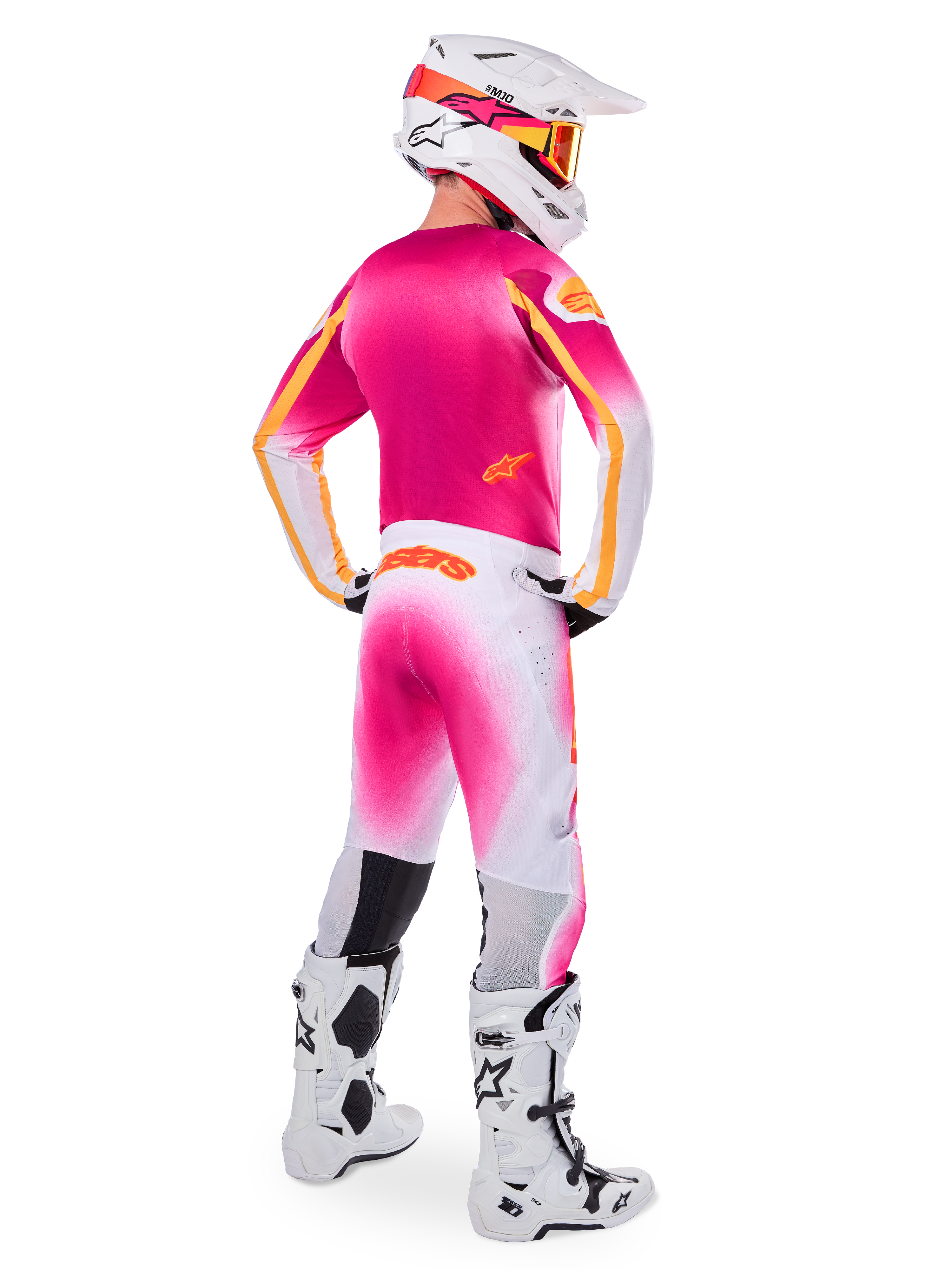 Pilote de motocross vu de dos portant la tenue Limited Edition Supertech AFD MIG, maillot rose avec accents orange, pantalon blanc avec dégradé rose et logo Alpinestars orange, casque blanc avec motifs roses et lunettes orange, bottes blanches et noires, équipement de course professionnel tout-terrain