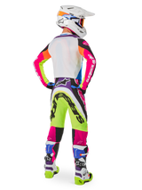 Pilote de motocross vu de dos portant l'équipement Alpinestars Limited Edition Supertech Hay, pantalon Supertech Hay multicolore aux graphismes jaune fluo, rose vif et violet, maillot assorti, casque blanc et bottes coordonnées blanches et bleues, tenue de course tout-terrain complète