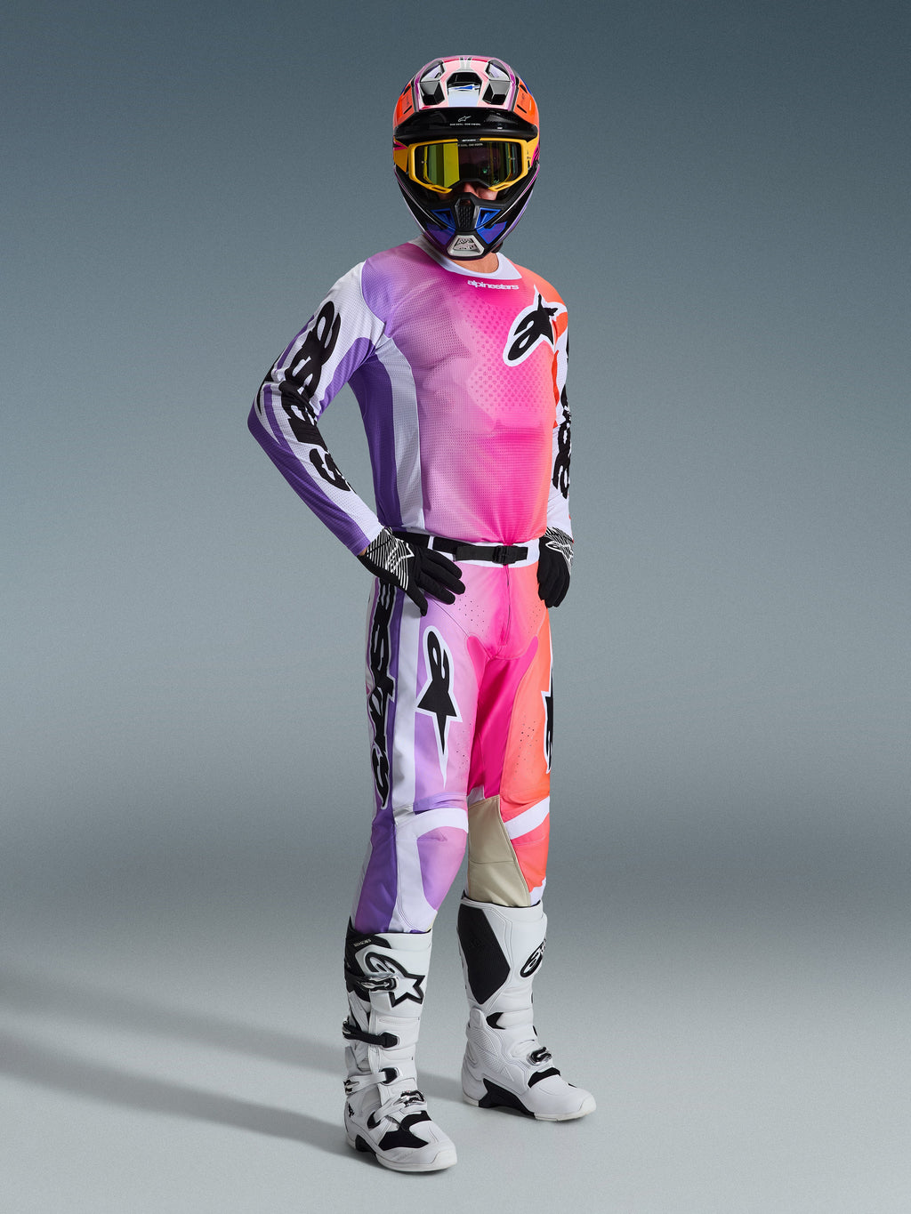 Pilote de motocross portant le maillot Alpinestars Racer Air Portl 2026 et son pantalon assorti, dégradé multicolore vif rose, violet et orange avec accents blancs, casque coloré coordonné et masque jaune, bottes blanches et noires, tenue de course complète pour la performance tout-terrain