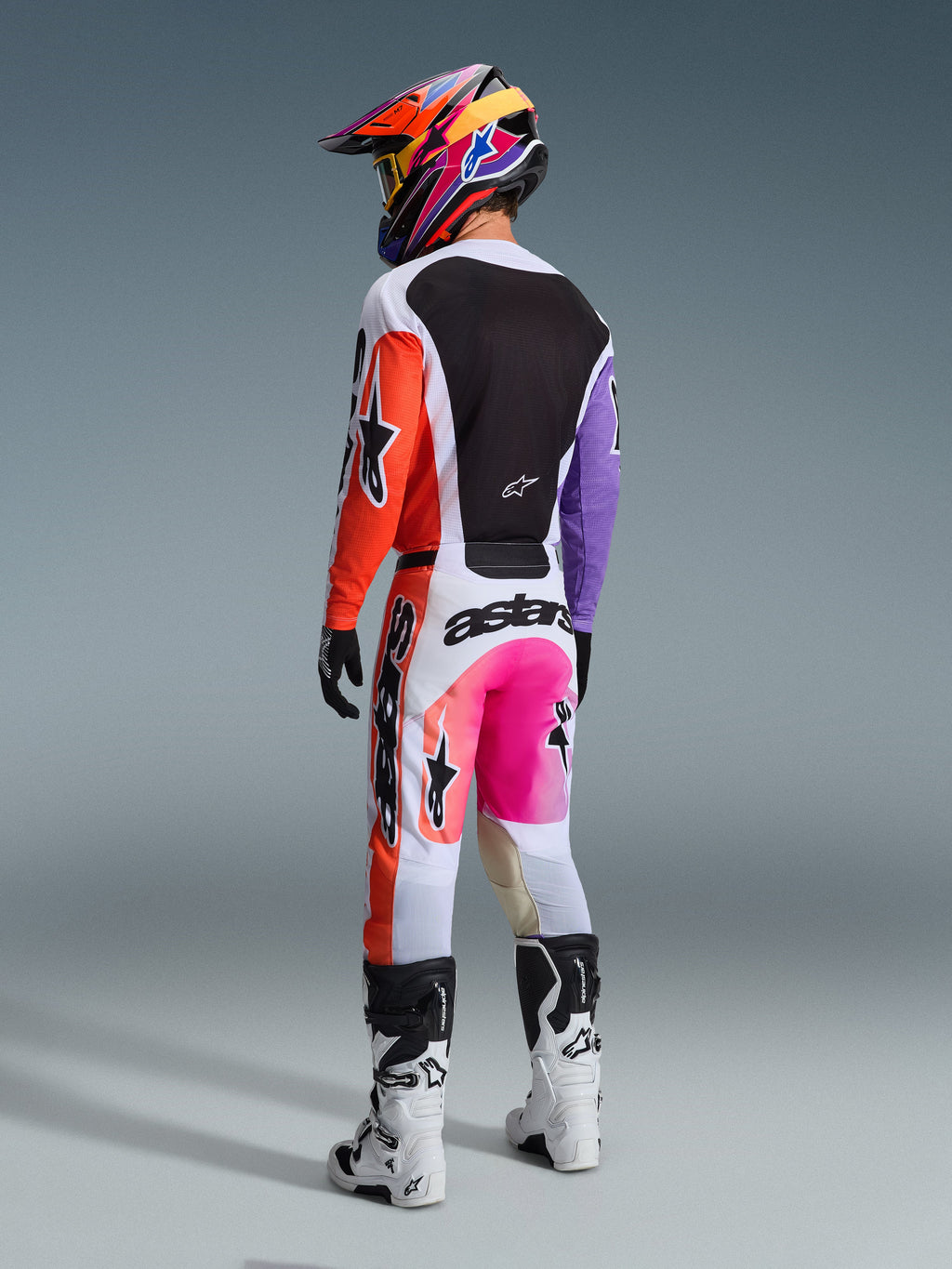 Maillot Alpinestars 2026 Racer Air Portl, maillot de motocross multicolore, vue arrière d'un pilote en tenue de course complète, présentant une base blanche avec des manches orange et violettes et un panneau dorsal en maille noire, associé à un pantalon rose et orange assorti, un casque coloré et des bottes blanches, tenue de course tout-terrain professionnelle