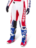 Alpinestars Limited Edition Techstar Fortitude Pants, pantalon de motocross, rouge vif, blanc et bleu, porté avec des bottes et gants tout-terrain assortis, logo vertical sur la jambe, panneaux perforés respirants et sections renforcées noires aux genoux, coupe de course athlétique