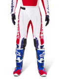 Alpinestars Limited Edition Techstar Fortitude Pants, pantalon de motocross, rouge vif, blanc et bleu, porté avec l'équipement assorti incluant des bottes de protection et des gants bleus, présentant un châssis blanc avec des graphismes rouges, des empiècements aux genoux en cuir noir et des perforations découpées au laser, tenue de course tout-terrain professionnelle