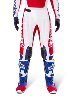 Alpinestars Limited Edition Techstar Fortitude Pants, pantalon de motocross, rouge vif, blanc et bleu, porté avec l'équipement assorti incluant des bottes de protection et des gants bleus, présentant un châssis blanc avec des graphismes rouges, des empiècements aux genoux en cuir noir et des perforations découpées au laser, tenue de course tout-terrain professionnelle