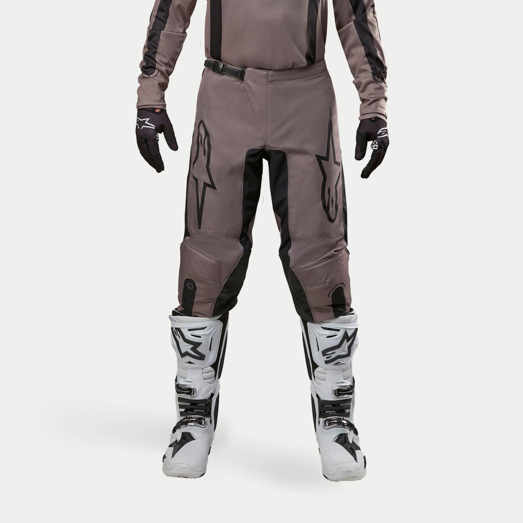 Pantalon Alpinestars Fluid Lurv 2024, pantalon de motocross, Mud Black, porté avec un maillot assorti, des gants noirs et des bottes blanches, avec de grands logos noirs sur les cuisses, une coupe ergonomique et un matériau durable pour la course tout-terrain