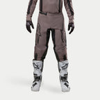 Pantalon Alpinestars Fluid Lurv 2024, pantalon de motocross, Mud Black, porté avec un maillot assorti, des gants noirs et des bottes blanches, avec de grands logos noirs sur les cuisses, une coupe ergonomique et un matériau durable pour la course tout-terrain