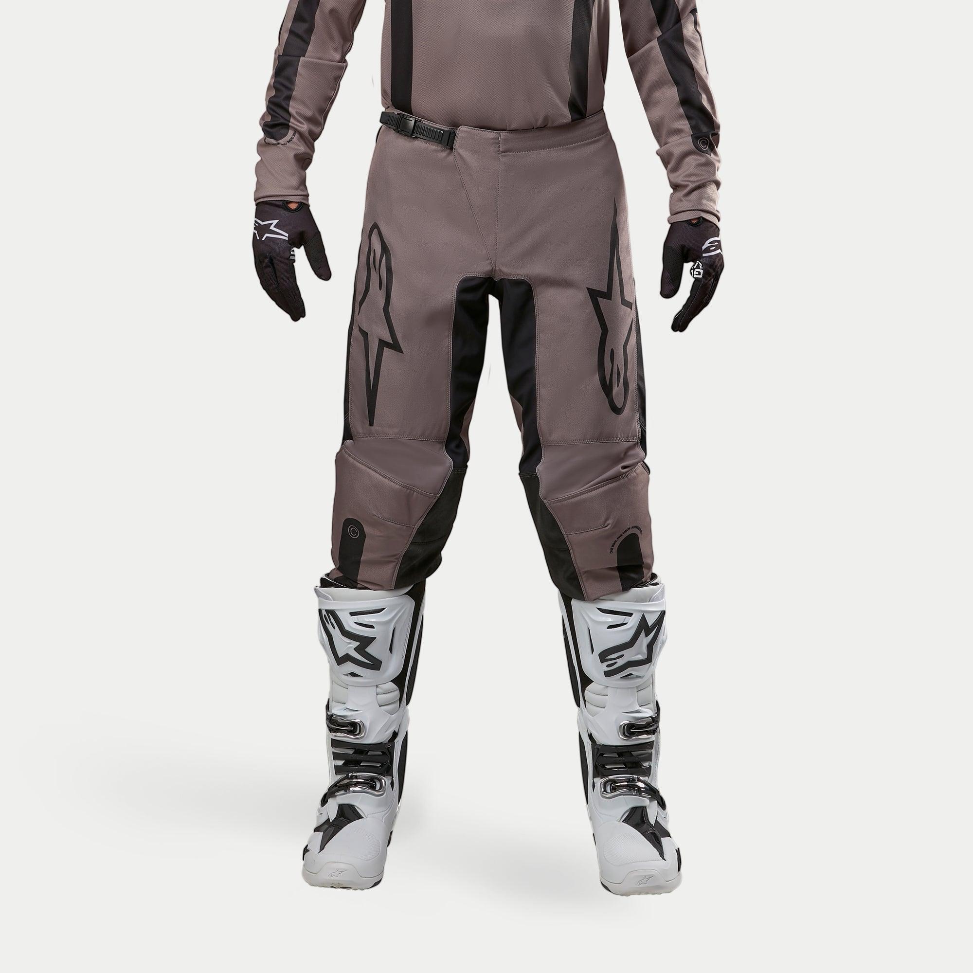 Pantalon Alpinestars Fluid Lurv 2024, pantalon de motocross, Mud Black, porté avec un maillot assorti, des gants noirs et des bottes blanches, avec de grands logos noirs sur les cuisses, une coupe ergonomique et un matériau durable pour la course tout-terrain