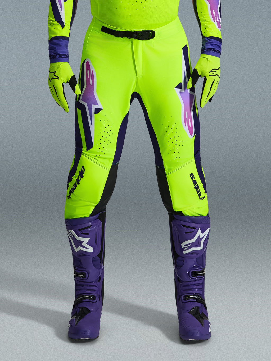 Pantalon Alpinestars Supertech Vista 2026, pantalon de motocross, jaune fluorescent, violet et noir, porté avec des gants assortis et des bottes violettes, présentant de grands logos en dégradé sur les cuisses, des zones perforées pour la ventilation et un design athlétique professionnel pour la course tout-terrain