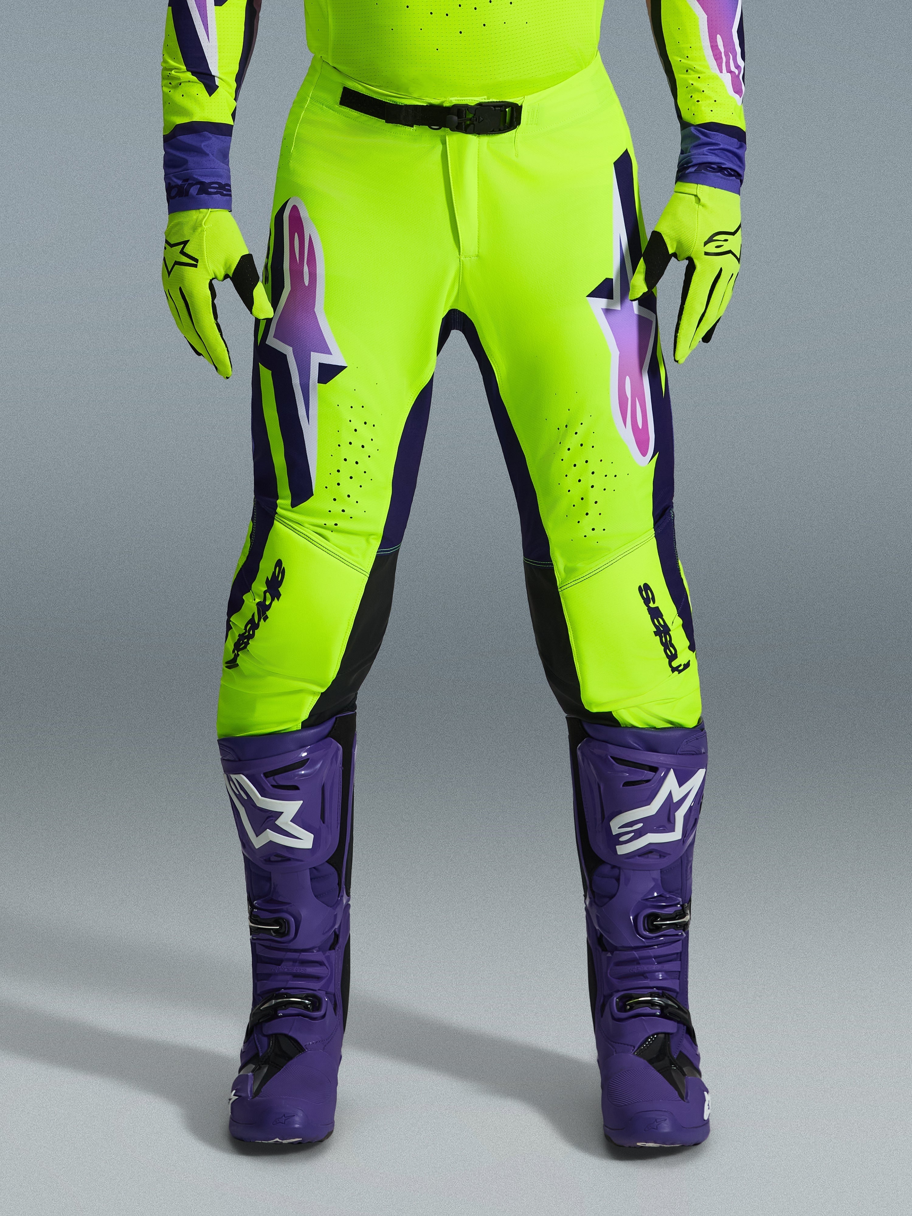 Pantalon Alpinestars Supertech Vista 2026, pantalon de motocross, jaune fluorescent, violet et noir, porté avec des gants assortis et des bottes violettes, présentant de grands logos en dégradé sur les cuisses, des zones perforées pour la ventilation et un design athlétique professionnel pour la course tout-terrain