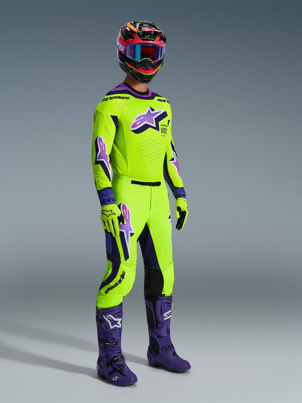Pantalon Alpinestars Supertech Vista 2026, pantalon de motocross, jaune néon et violet, porté avec un maillot de course assorti, des bottes violettes et un casque coloré, doté de panneaux en cuir noir aux genoux, de logos Astar violets et de zones de ventilation perforées, tenue de course tout-terrain coordonnée.