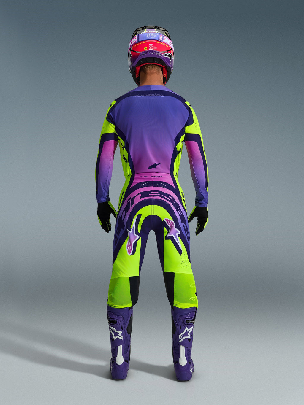 Pilote de motocross vu de dos portant le maillot Supertech Vista 2026 et le pantalon assorti, design jaune et violet éclatant avec dégradés roses, bottes violettes avec logos Alpinestars blancs, gants noirs et casque de course rouge et violet, ensemble d'équipement de course professionnel.