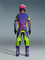 Pilote de motocross vu de dos portant le maillot Supertech Vista 2026 et le pantalon assorti, design jaune et violet éclatant avec dégradés roses, bottes violettes avec logos Alpinestars blancs, gants noirs et casque de course rouge et violet, ensemble d'équipement de course professionnel.
