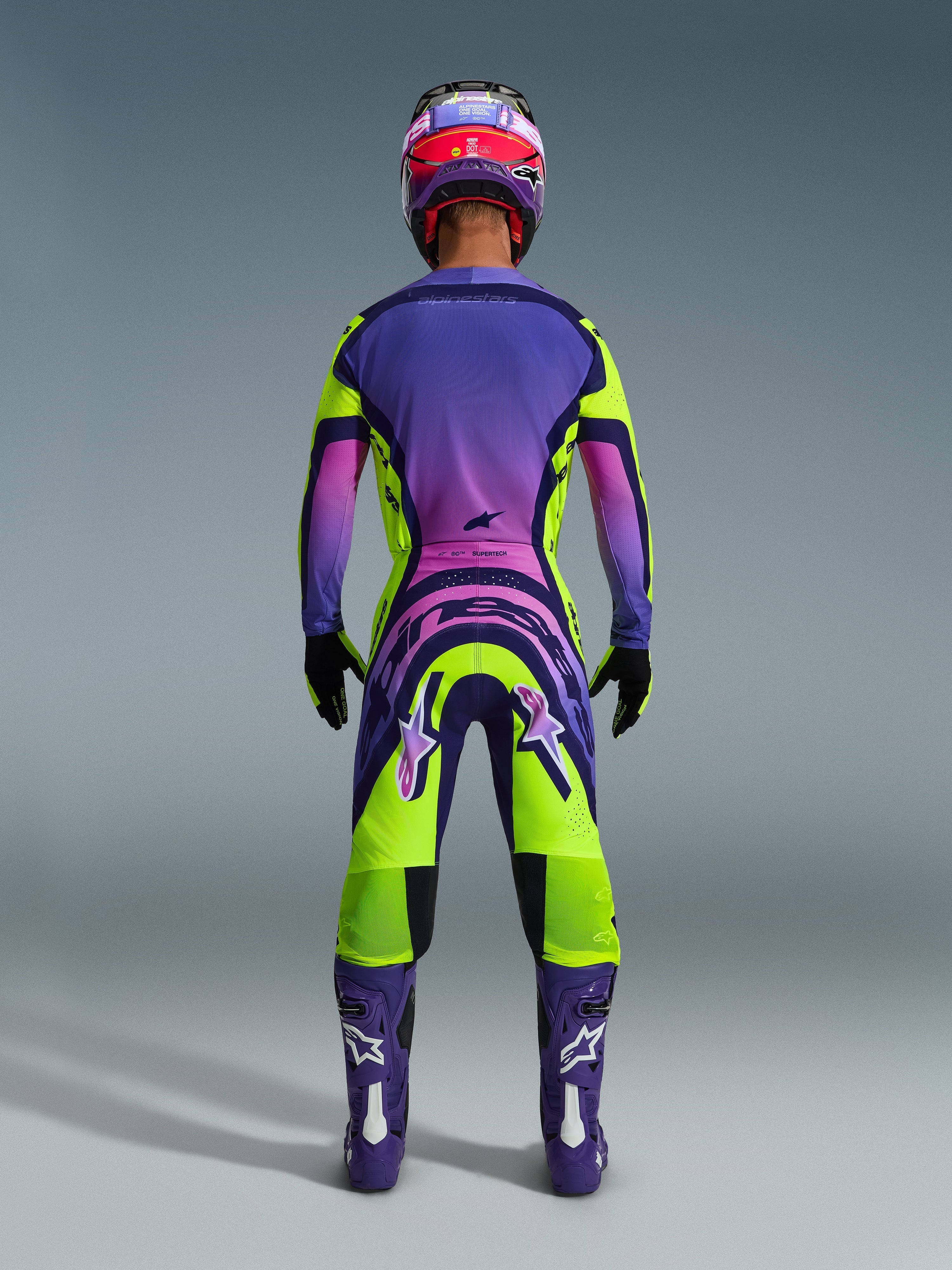 Pilote de motocross vu de dos portant le maillot Supertech Vista 2026 et le pantalon assorti, design jaune et violet éclatant avec dégradés roses, bottes violettes avec logos Alpinestars blancs, gants noirs et casque de course rouge et violet, ensemble d'équipement de course professionnel.