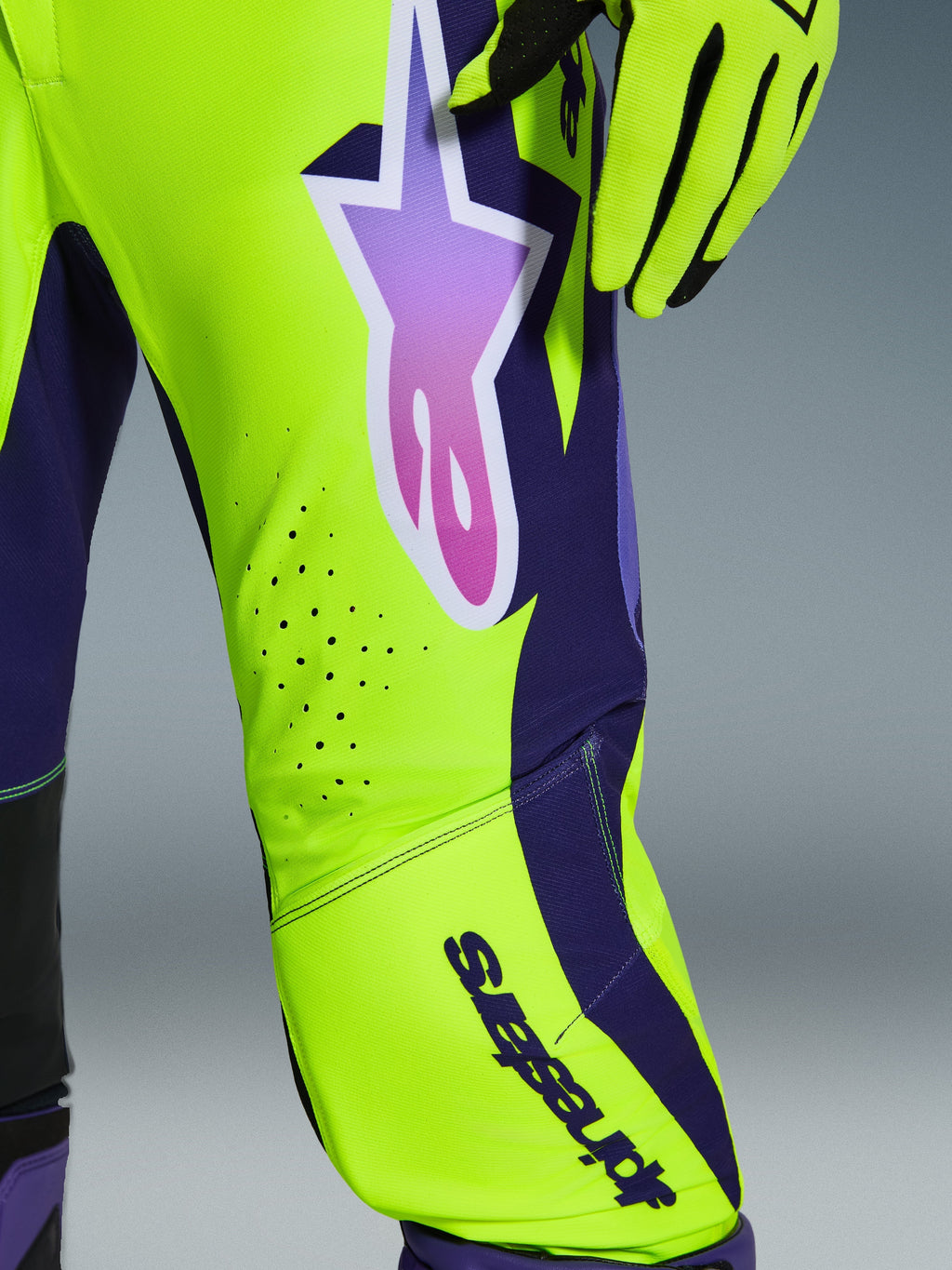 Pantalon Alpinestars Supertech Vista 2026, pantalon de motocross, jaune, violet et noir, gros plan montrant le logo étoile en dégradé et les perforations de ventilation découpées au laser, associé à des gants de course assortis