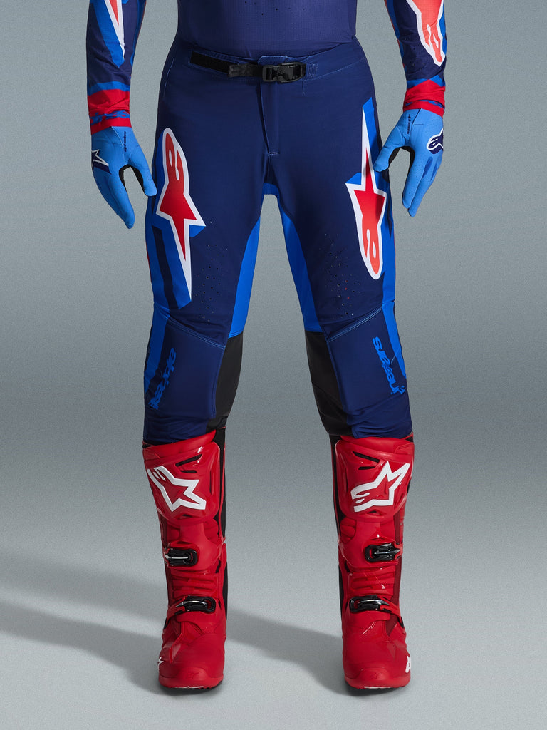Alpinestars 2026 Supertech Vista Pants, pantalon de motocross, bleu marine foncé, corail vif et cobalt, vue de face d'un pilote portant une tenue de performance avec des gants bleus et des bottes de motocross rouges assortis, arborant des logos Alpinestars rouges et blancs sur les cuisses.