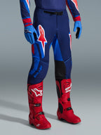Alpinestars Pantalon Supertech Vista 2026, pantalon de motocross, bleu marine foncé, corail vif et bleu cobalt, porté avec des bottes de course rouge vif et des gants bleus, avec de grands logos graphiques sur les cuisses et des panneaux intérieurs de genoux renforcés pour la performance tout-terrain