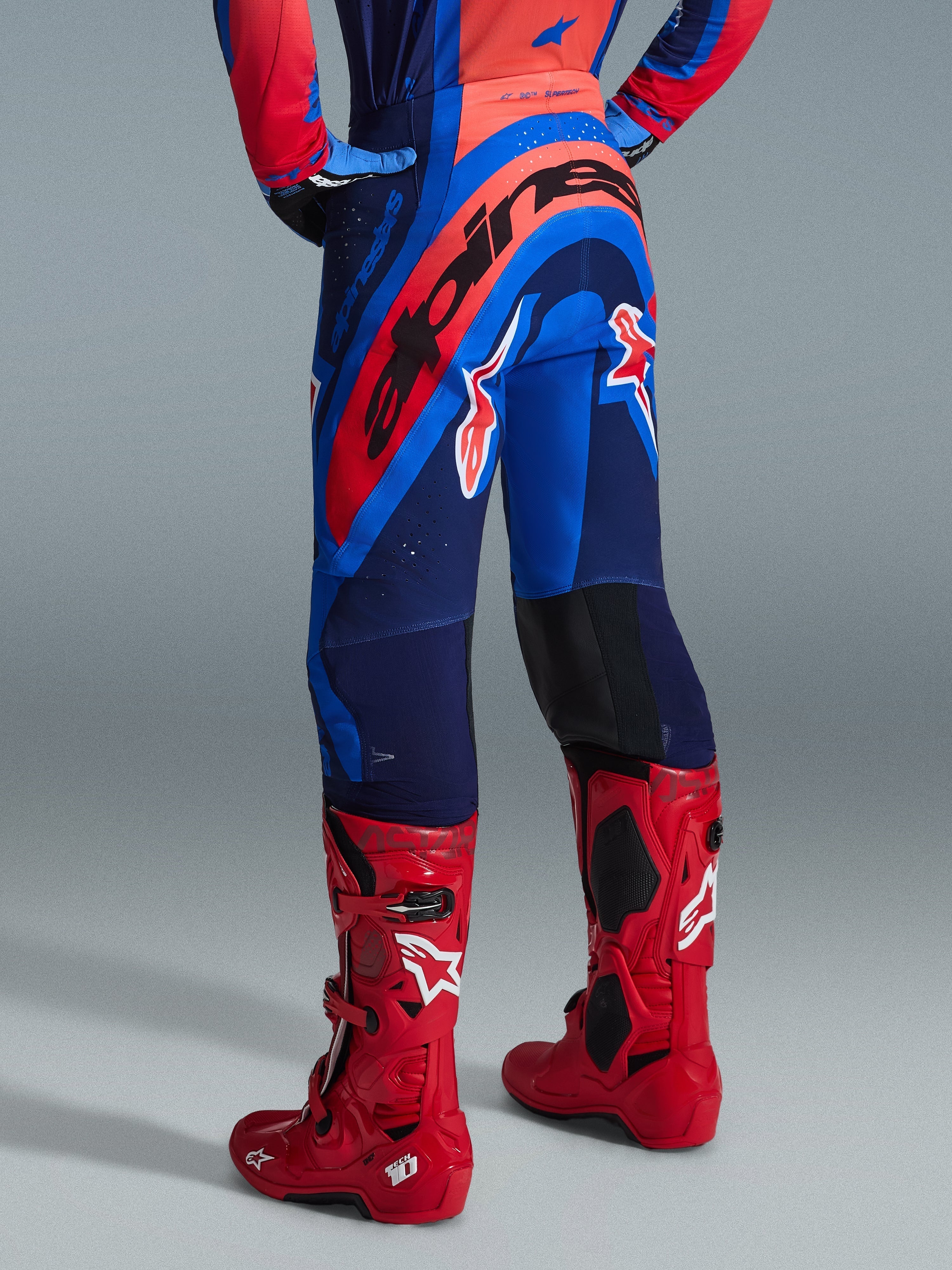 Pantalon Alpinestars Supertech Vista 2026, pantalon de motocross, bleu marine foncé, corail éclatant et cobalt, vue arrière du pilote, associé à des bottes de motocross rouges, marquage imposant sur l'assise, coupe athlétique avec zones extensibles et construction rip-stop durable pour la compétition professionnelle