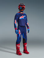 Pilote de motocross portant le pantalon Supertech Vista 2026 en coloris Dark Navy Hot Coral Cobalt, maillot de course à manches longues assorti, bottes de motocross rouges et casque coordonné avec lunettes rouges, prise de vue en studio plein pied d'un équipement tout-terrain haute performance