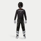 Pilote de motocross portant une tenue de course complète, pantalon Alpinestars 2024 Fluid Graphite noir et argent, maillot noir à manches longues avec logo argenté, casque noir avec lunettes, bottes blanches et noires, gants blancs et noirs, tenue de motocross coordonnée