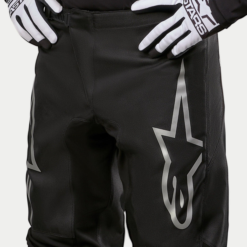 Alpinestars 2024 Fluid Graphite Pants, pantalon de motocross, noir et argent, gros plan sur le haut des cuisses et la taille, avec un grand logo étoile argenté sur la jambe, porté avec des gants de course noirs et blancs