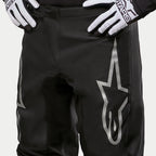 Alpinestars 2024 Fluid Graphite Pants, pantalon de motocross, noir et argent, gros plan sur le haut des cuisses et la taille, avec un grand logo étoile argenté sur la jambe, porté avec des gants de course noirs et blancs