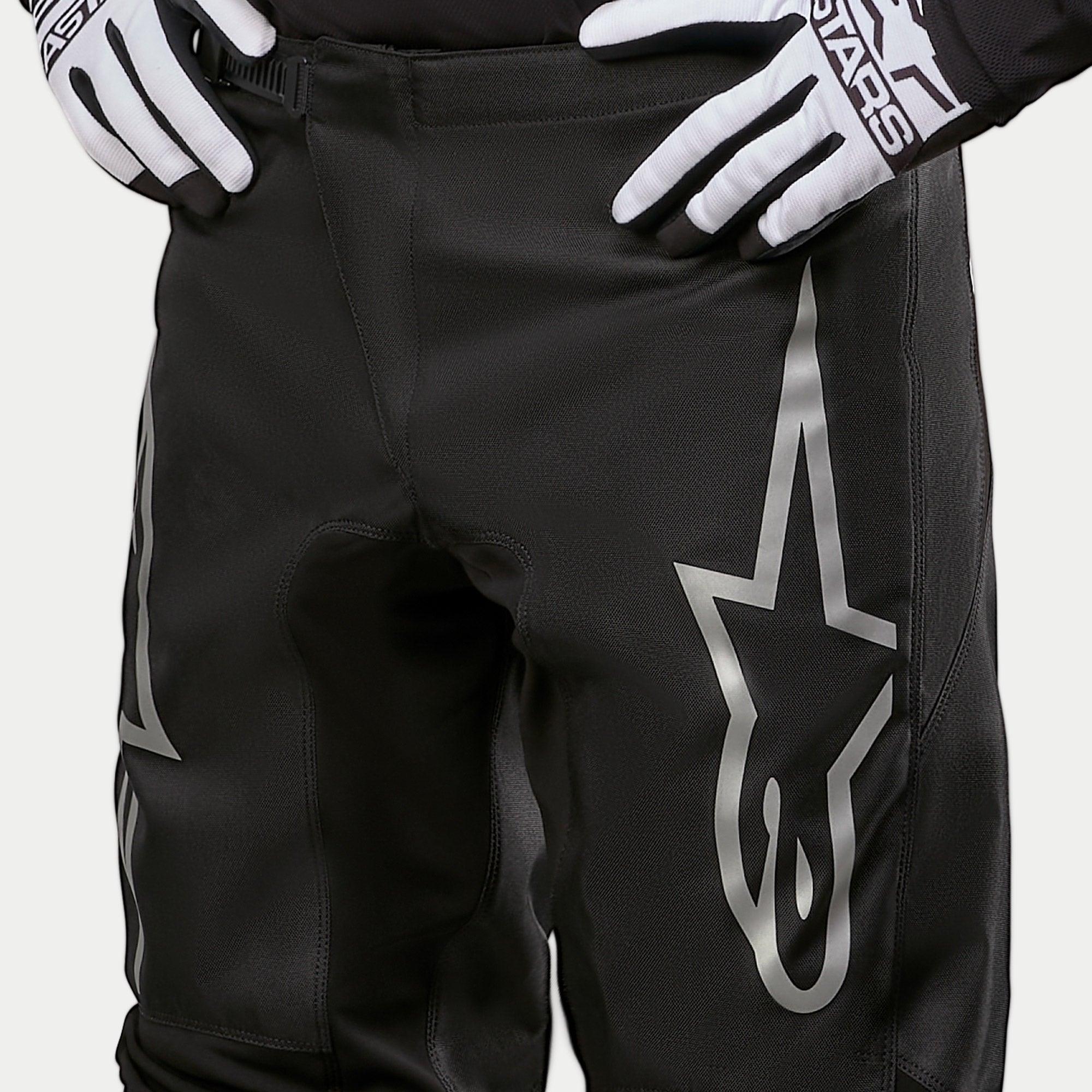 Alpinestars 2024 Fluid Graphite Pants, pantalon de motocross, noir et argent, gros plan sur le haut des cuisses et la taille, avec un grand logo étoile argenté sur la jambe, porté avec des gants de course noirs et blancs