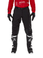 Pantalon Alpinestars Techdura, pantalon d'enduro et de tout-terrain, noir, porté avec un maillot Alpinestars rouge et des bottes noires et blanches, doté d'une construction en toile extensible robuste, d'une boucle de ceinture magnétique réglable et de renforts de genoux en cuir pour la performance tout-terrain