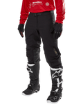 Alpinestars Techdura Pants, pantalon de moto tout-terrain noir, porté avec un maillot rouge à manches longues et des bottes de motocross noires et blanches, doté d'une construction robuste en polyamide extensible, de renforts de genoux internes en cuir et d'une ceinture à boucle réglable pour l'enduro.