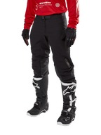 Alpinestars Techdura Pants, pantalon de moto tout-terrain noir, porté avec un maillot rouge à manches longues et des bottes de motocross noires et blanches, doté d'une construction robuste en polyamide extensible, de renforts de genoux internes en cuir et d'une ceinture à boucle réglable pour l'enduro.