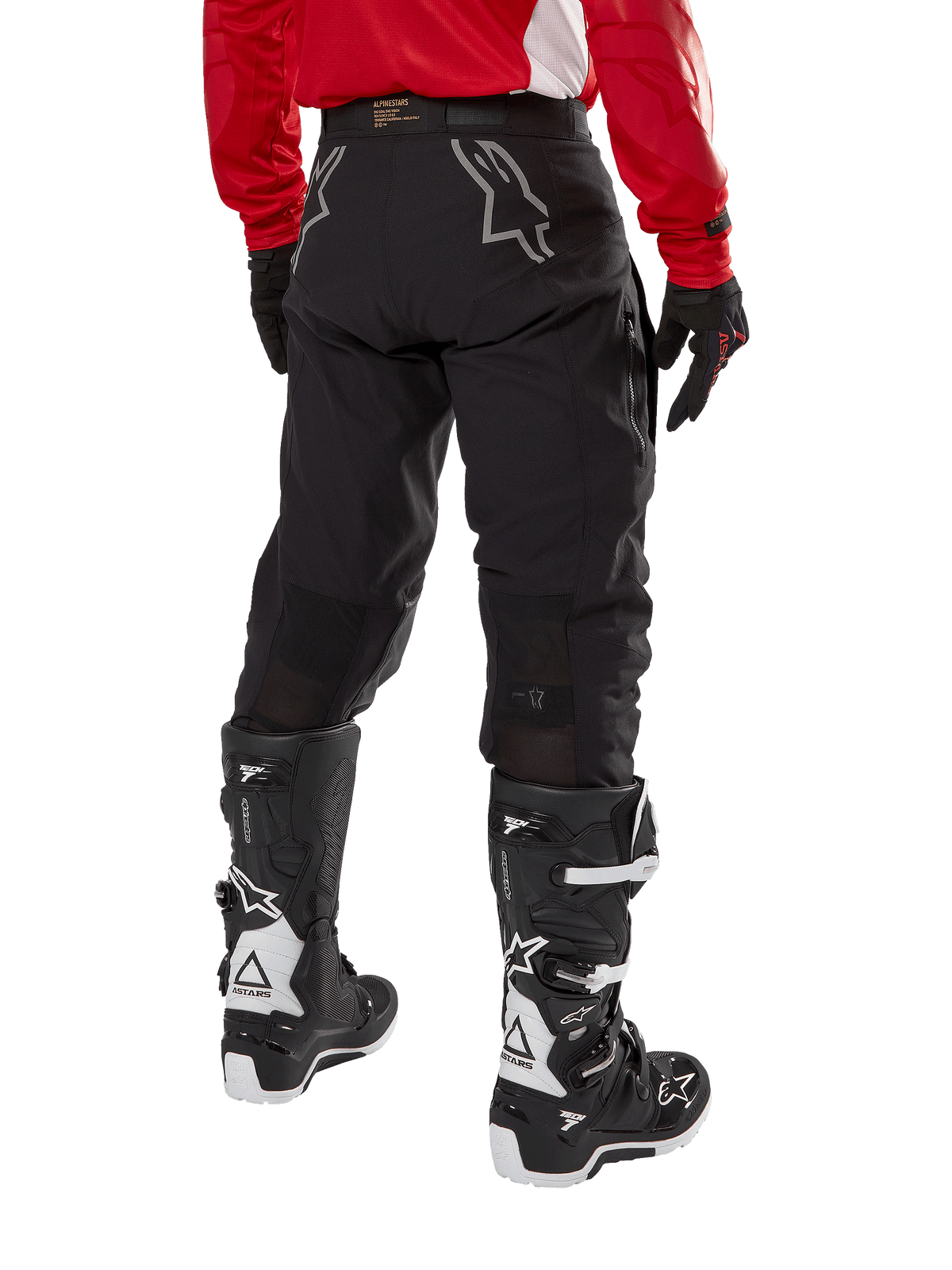 Pantalon Alpinestars Techdura, pantalon d'enduro tout-terrain, noir, vue arrière montrant des logos d'étoile gris sur l'assise, porté avec un maillot rouge à manches longues et des bottes Tech 7 noires et blanches, construction textile robuste pour le pilotage d'aventure