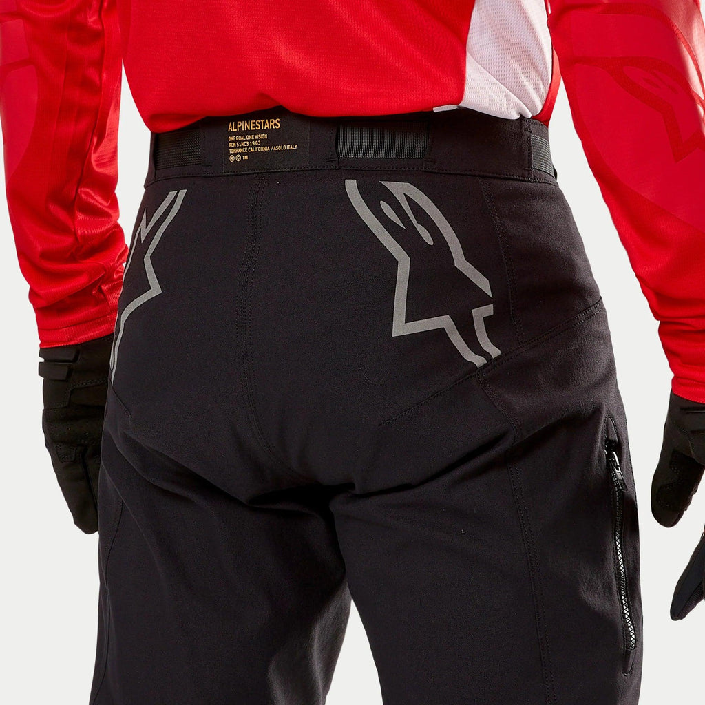 Alpinestars Techdura Pants, pantalon d'enduro noir, vue arrière avec de grands logos Astars gris sur l'assise, porté avec un maillot rouge et des gants noirs, matériau robuste avec fermeture éclair latérale et ceinture logotypée