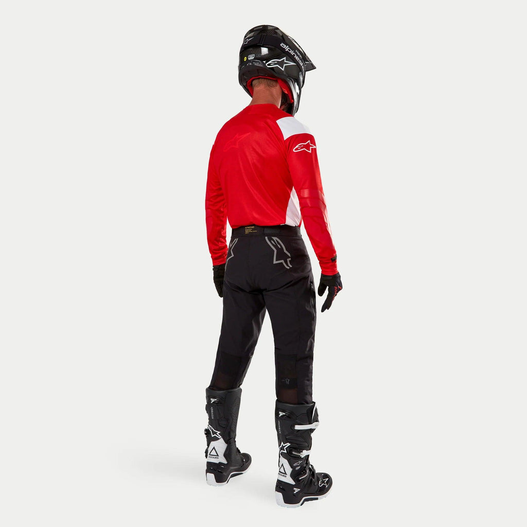 Alpinestars Techdura Pants, pantalon d'enduro tout-terrain, noir, vue arrière porté par un pilote avec un maillot rouge et blanc et des bottes noires et blanches, doté de logos d'étoiles blancs au dos et d'une construction technique extensible et durable.