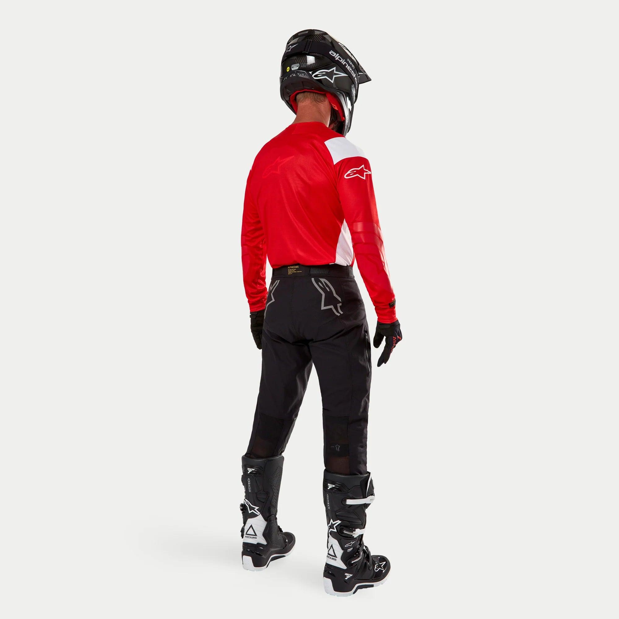Alpinestars Techdura Pants, pantalon d'enduro tout-terrain, noir, vue arrière porté par un pilote avec un maillot rouge et blanc et des bottes noires et blanches, doté de logos d'étoiles blancs au dos et d'une construction technique extensible et durable.