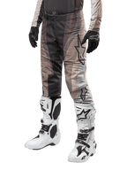 Pantalon Alpinestars Techstar Pneuma 2024, pantalon de motocross, sable foncé et gris poussière de fer, porté avec des bottes blanches et noires et des gants noirs, avec un motif ondulé et un grand logo étoile, équipement durable pour la course tout-terrain
