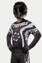 Alpinestars 2024 Racer Hana Jersey, maillot de motocross, noir et blanc, avec des graphismes et un logo Alpinestars blancs audacieux, porté par un pilote avec un casque et des lunettes noirs, équipement haute performance pour la course tout-terrain