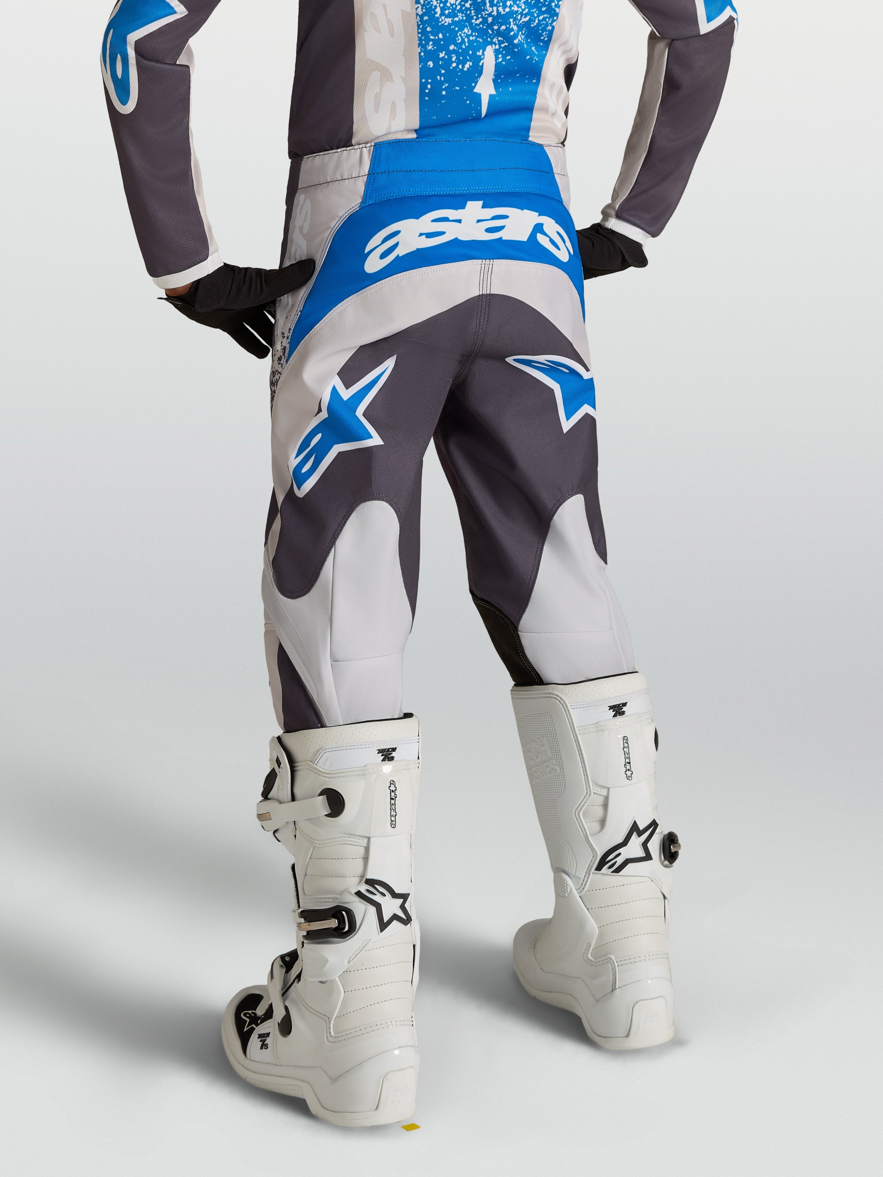Pantalon Fluid Drag Junior 2027 d'Alpinestars, pantalon de motocross gris et bleu pour enfant, vu de dos avec des bottes de moto blanches.