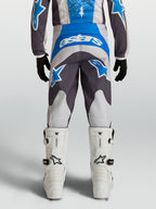 Alpinestars Pantalon Fluid Drag Junior, pantalon de motocross tout-terrain, bleu et gris, vue de dos montrant la coupe préformée, porté avec des bottes de moto blanches.