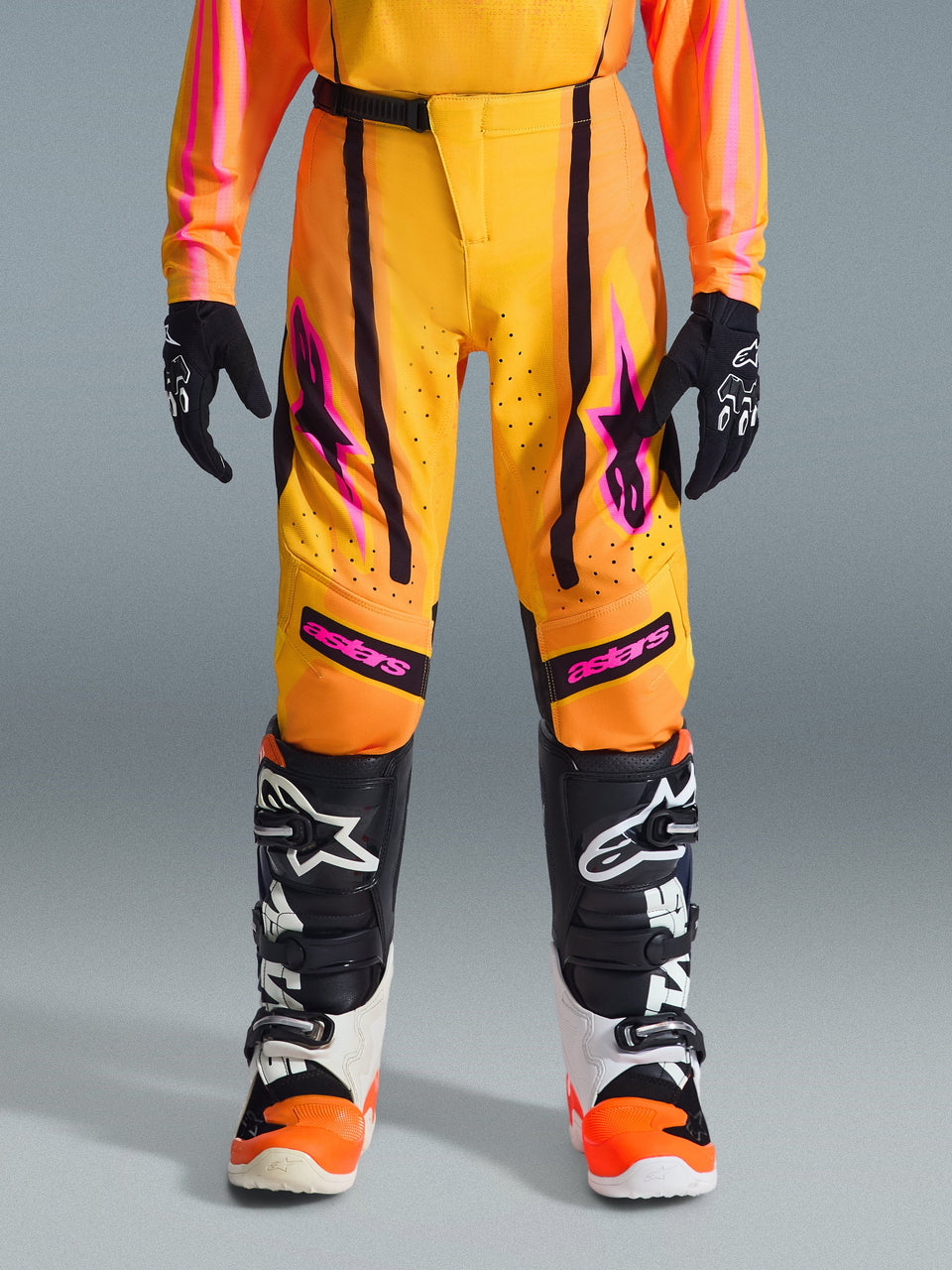 Pantalon Alpinestars Youth Supertech Nomur 2026, pantalon de motocross, orange, jaune et rose, porté avec un maillot et des bottes assortis, avec des rayures noires, des logos étoiles roses et des zones perforées, conçu pour la compétition de motocross junior