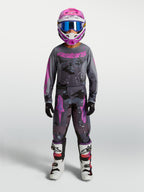 Pantalon Fluid Lone Junior Alpinestars, équipement de motocross pour enfant, gris camouflage et violet, porté par un pilote avec maillot assorti et bottes blanches, construction ergonomique en polyfibre 600D.