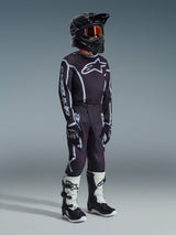 Alpinestars 2026 Youth Fluid Apex Pants, pantalon de motocross, noir et gris, porté avec un maillot noir et blanc assorti, un casque noir et des bottes blanches et noires, arborant un logo Alpinestars audacieux et des lignes graphiques, tenue de course coordonnée pour le motocross junior